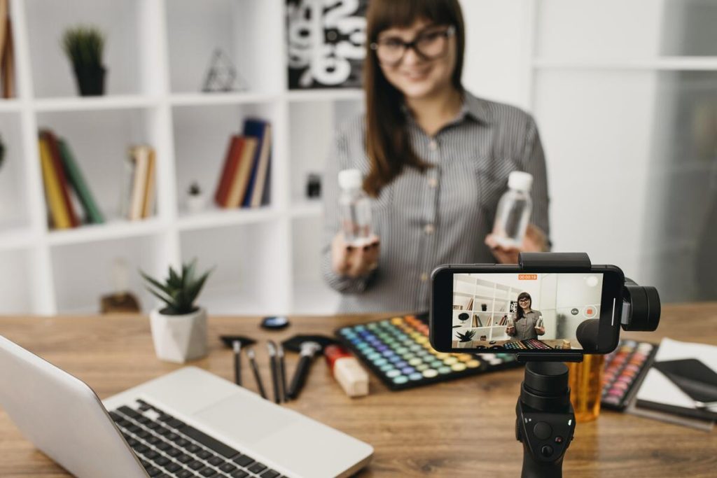 Mulher sorridente gravando conteúdo com celular apoiado em um tripé, segurando dois frascos e rodeada por itens de maquiagem sobre a mesa, representando uma estratégia de vídeo marketing.